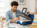 Hombre sonriente usando una plancha de calor para un ejemplo de sublimación en una camiseta. Hay tazas y cojines con diseños coloridos.