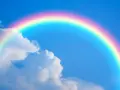 Un arcoíris con sus vibrantes colores del arcoiris cruza un cielo azul claro con nubes blancas.