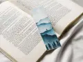 Un marcapáginas con un paisaje montañoso y árboles en acuarela, colocado sobre un libro abierto.