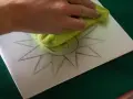 Mano limpiando una superficie metálica con un paño verde, sobre la cual se dibuja un sol. Preparando una litografía casera.