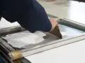 Manos aplicando tinta blanca con rasqueta sobre pantalla para serigrafía en casa.