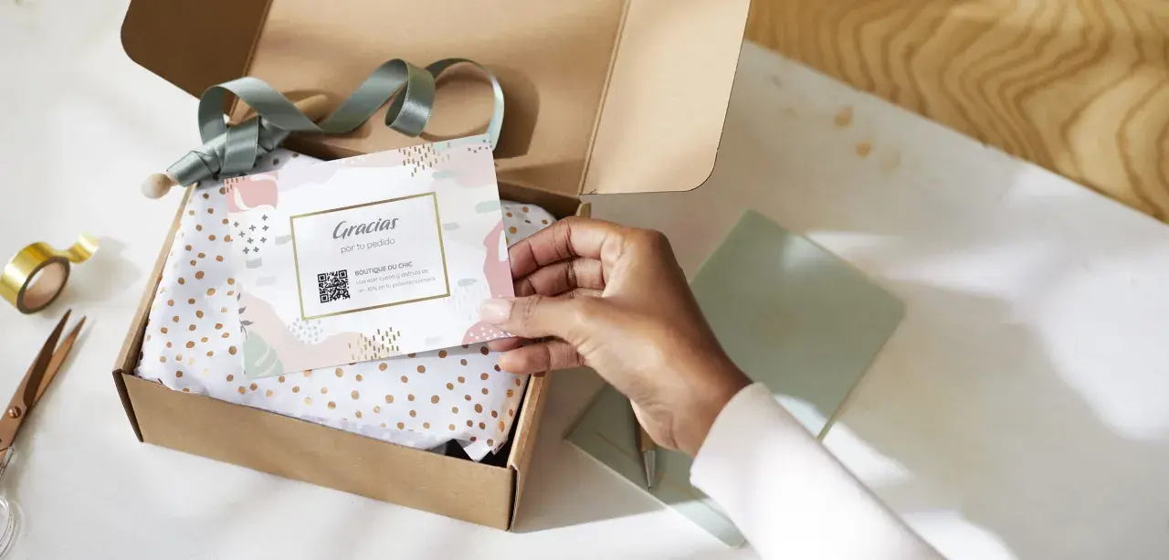 Tarjeta de agradecimiento tamaño postal con código QR, dentro de una caja abierta con papel de seda dorado.