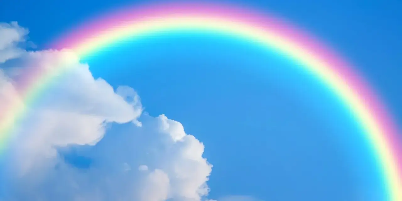 Un arcoíris con sus vibrantes colores del arcoiris cruza un cielo azul claro con nubes blancas.