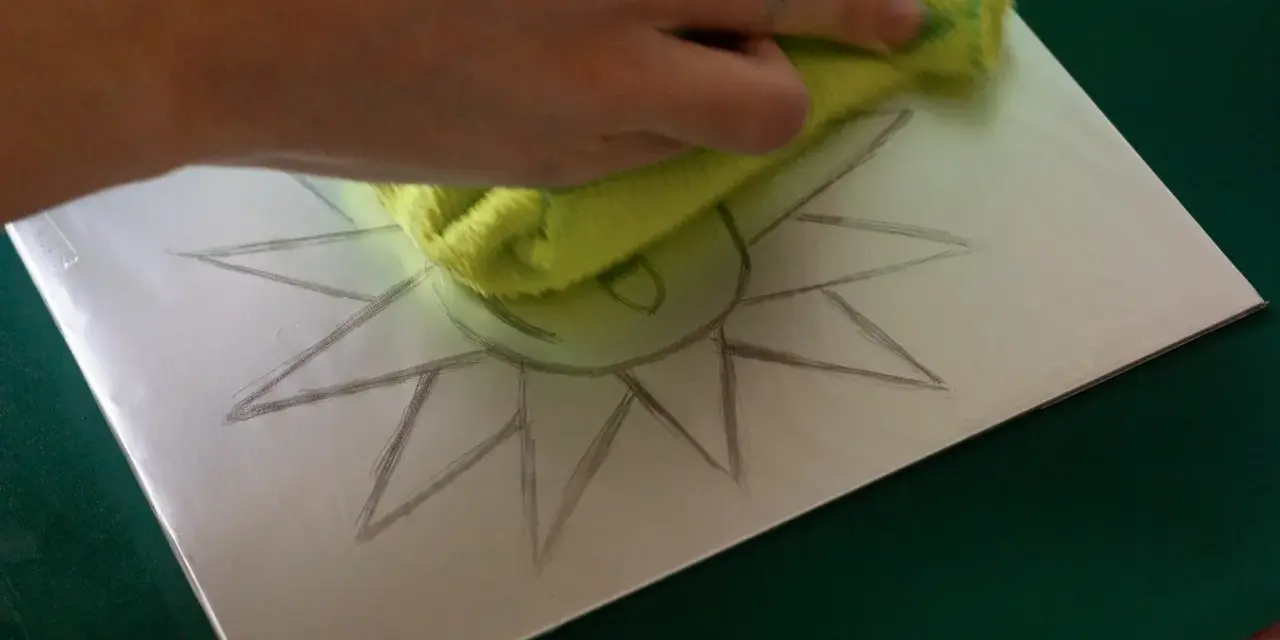Mano limpiando una superficie metálica con un paño verde, sobre la cual se dibuja un sol. Preparando una litografía casera.