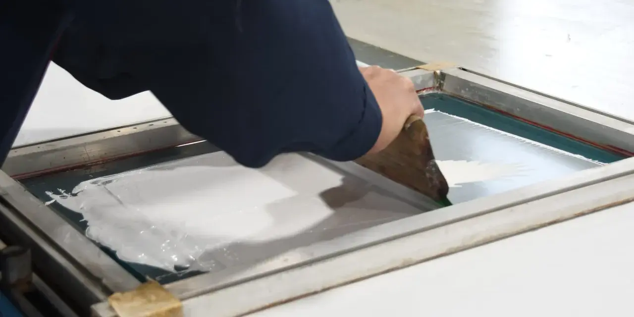 Manos aplicando tinta blanca con rasqueta sobre pantalla para serigrafía en casa.