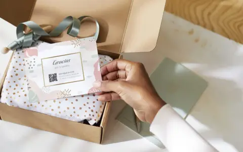 Tarjeta de agradecimiento tamaño postal con código QR, dentro de una caja abierta con papel de seda dorado.