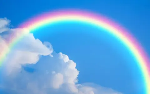 Un arcoíris con sus vibrantes colores del arcoiris cruza un cielo azul claro con nubes blancas.