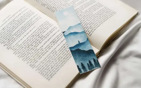 Un marcapáginas con un paisaje montañoso y árboles en acuarela, colocado sobre un libro abierto.