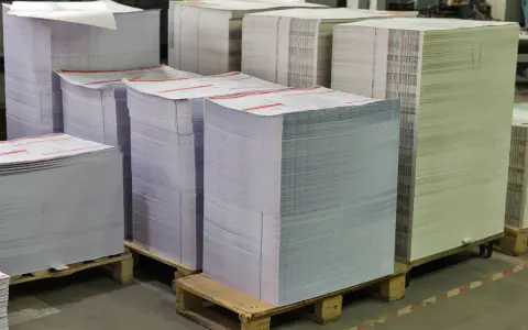 Grandes pilas de papel apiladas en palets, listas para ser impresas. Las medidas folio son evidentes en la cantidad de material.