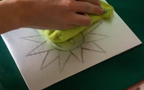 Mano limpiando una superficie metálica con un paño verde, sobre la cual se dibuja un sol. Preparando una litografía casera.
