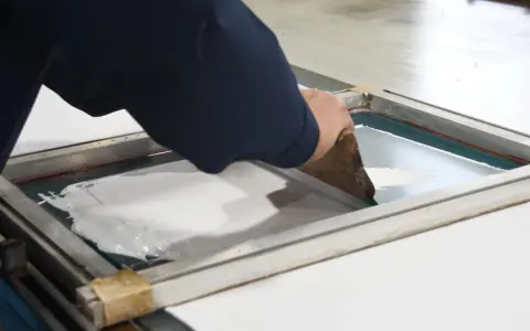 Manos aplicando tinta blanca con rasqueta sobre pantalla para serigrafía en casa.
