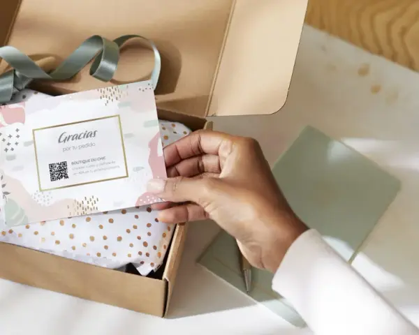 Tarjeta de agradecimiento tamaño postal con código QR, dentro de una caja abierta con papel de seda dorado.