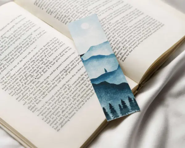 Un marcapáginas con un paisaje montañoso y árboles en acuarela, colocado sobre un libro abierto.
