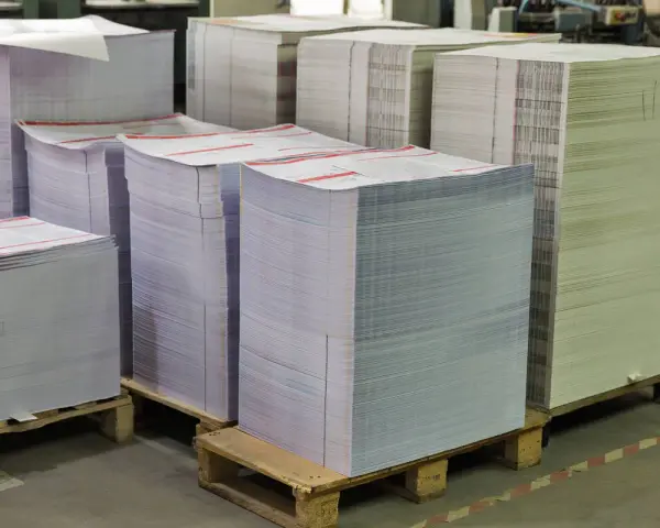 Grandes pilas de papel apiladas en palets, listas para ser impresas. Las medidas folio son evidentes en la cantidad de material.