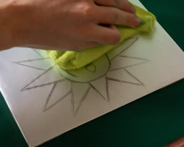Mano limpiando una superficie metálica con un paño verde, sobre la cual se dibuja un sol. Preparando una litografía casera.