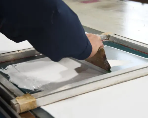 Manos aplicando tinta blanca con rasqueta sobre pantalla para serigrafía en casa.