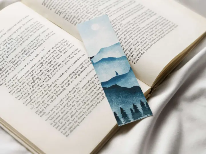 Un marcapáginas con un paisaje montañoso y árboles en acuarela, colocado sobre un libro abierto.