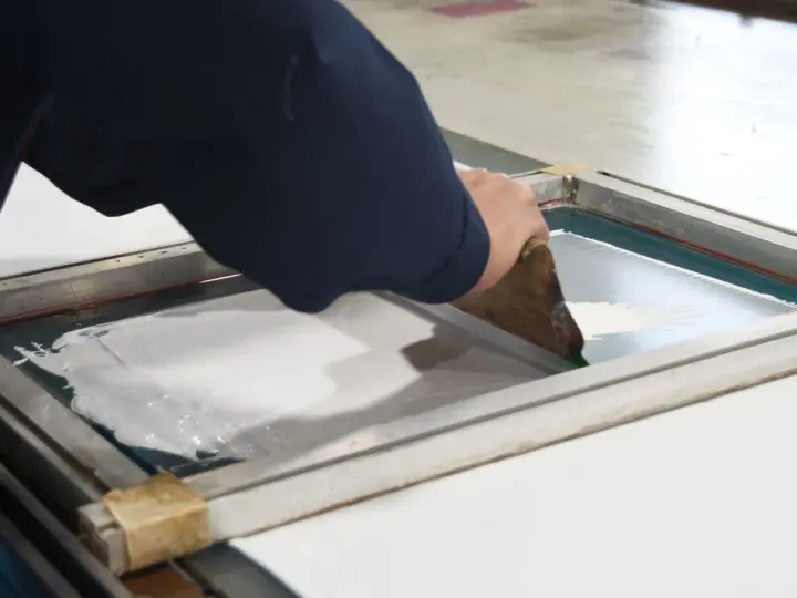 Manos aplicando tinta blanca con rasqueta sobre pantalla para serigrafía en casa.