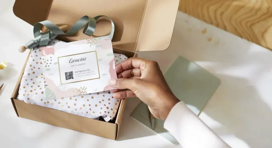 Tarjeta de agradecimiento tamaño postal con código QR, dentro de una caja abierta con papel de seda dorado.