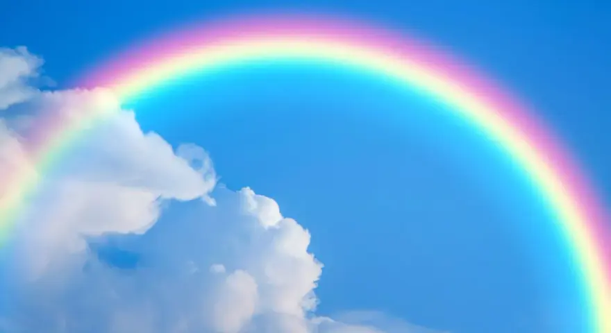 Un arcoíris con sus vibrantes colores del arcoiris cruza un cielo azul claro con nubes blancas.