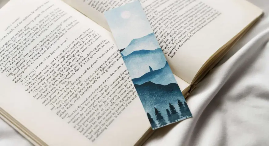 Un marcapáginas con un paisaje montañoso y árboles en acuarela, colocado sobre un libro abierto.