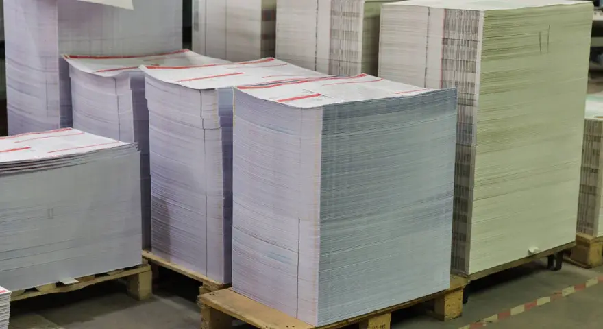 Grandes pilas de papel apiladas en palets, listas para ser impresas. Las medidas folio son evidentes en la cantidad de material.