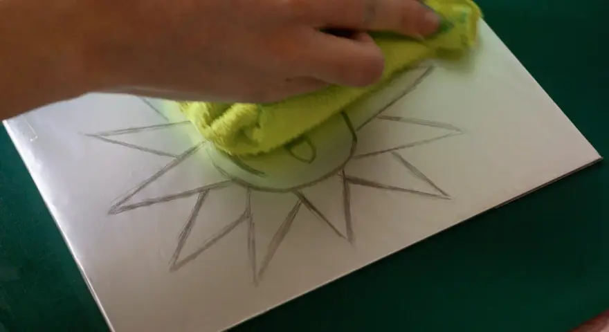 Mano limpiando una superficie metálica con un paño verde, sobre la cual se dibuja un sol. Preparando una litografía casera.
