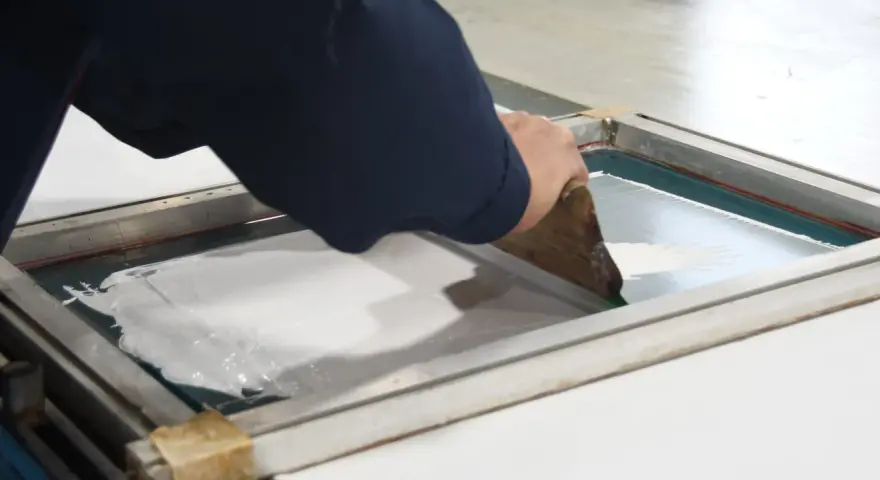 Manos aplicando tinta blanca con rasqueta sobre pantalla para serigrafía en casa.