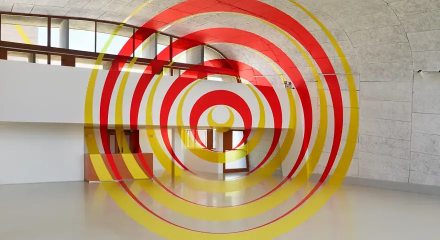 Círculos concéntricos rojos y amarillos que parecen flotar en un espacio arquitectónico moderno. Ejemplos de anamorfosis que transforman la percepción.