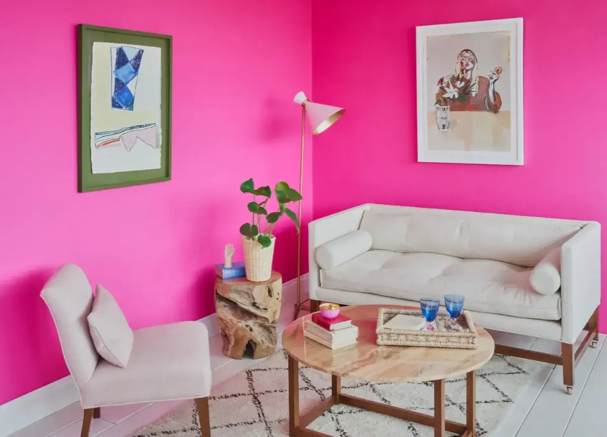 Un salón con paredes de el rosa mas rosa del mundo. Un sofá blanco, una silla rosa pálido, mesa de centro y arte abstracto.