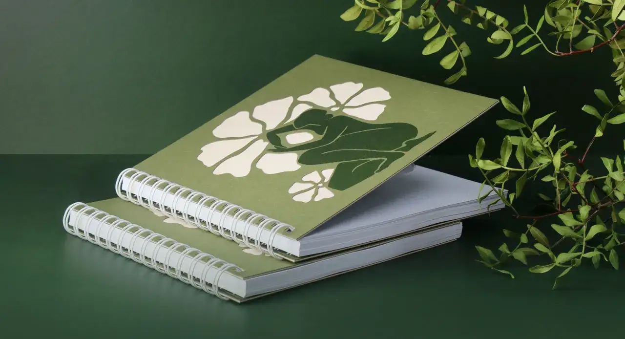 Dos cuadernos en espiral con una figura verde y flores blancas. La impresi&oacute;n gr&aacute;fica evoca la naturaleza.