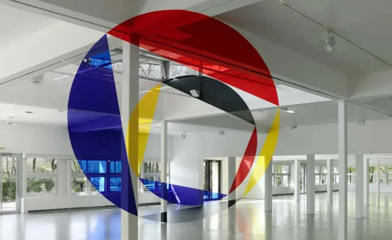 Instalaci&oacute;n art&iacute;stica con formas geom&eacute;tricas de colores (rojo, azul, amarillo) superpuestas en un espacio blanco, creando efectos de anamorfosis.