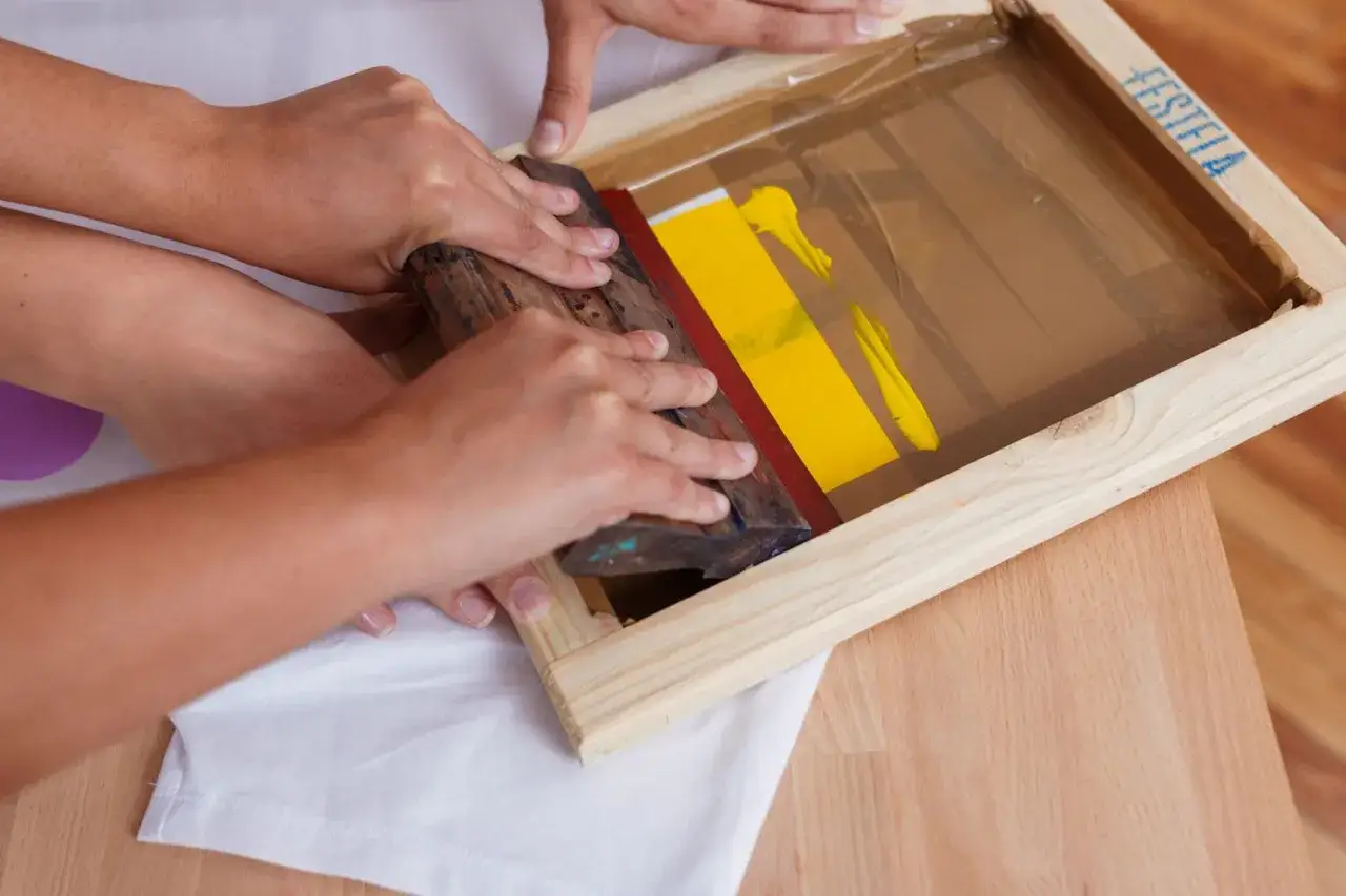 Manos aplicando tinta amarilla con rasqueta sobre pantalla para serigraf&iacute;a en casa.