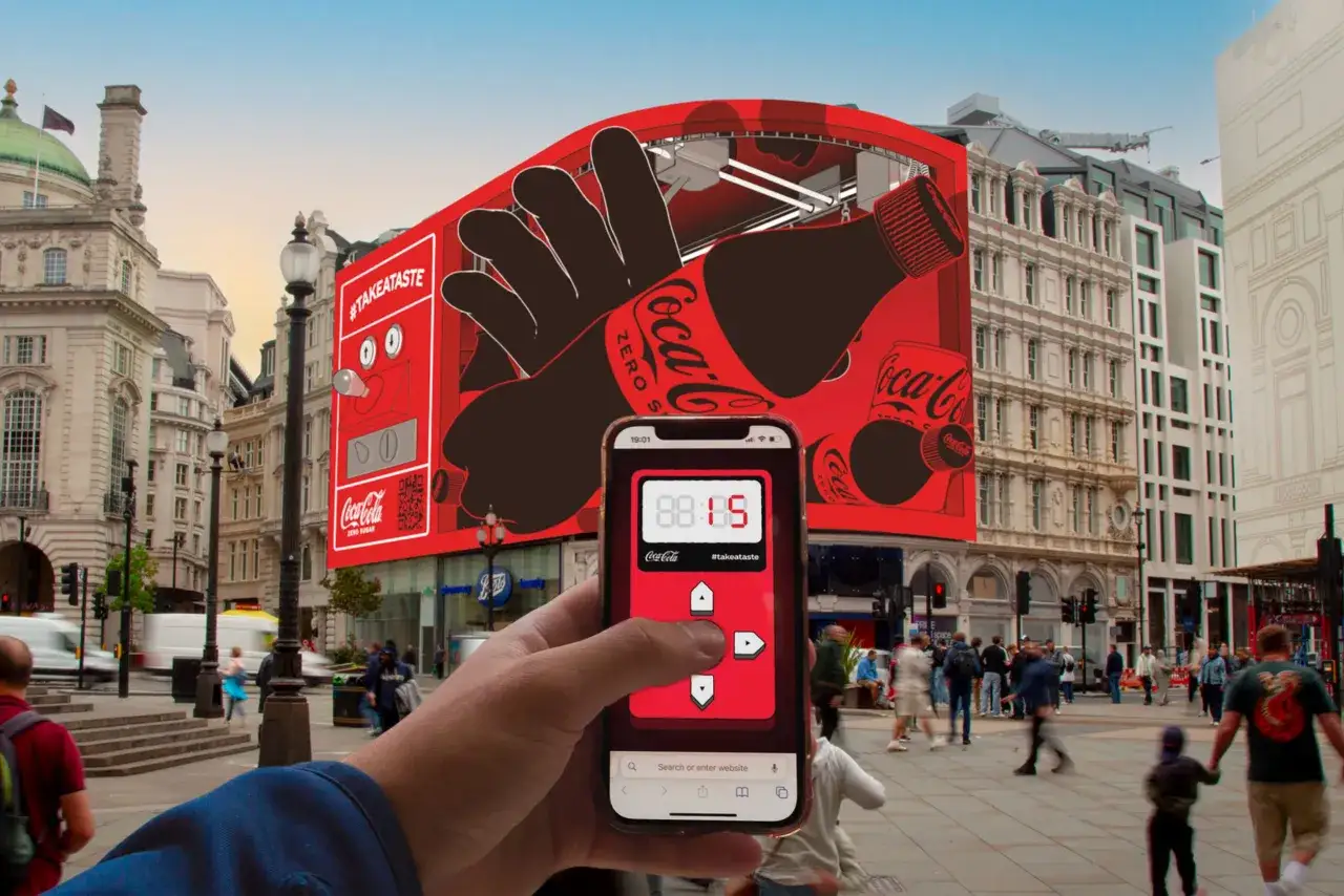 Una mano sostiene un teléfono con una interfaz de Coca-Cola Zero. Detrás, una pantalla gigante muestra una botella de Coca-Cola y el hashtag #takeataste, creando una experiencia de