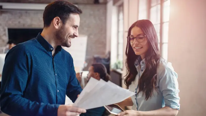 Hombre y mujer sonríen mientras revisan documentos, un ejemplo de cómo la publicidad social puede unir a las personas en el trabajo.
